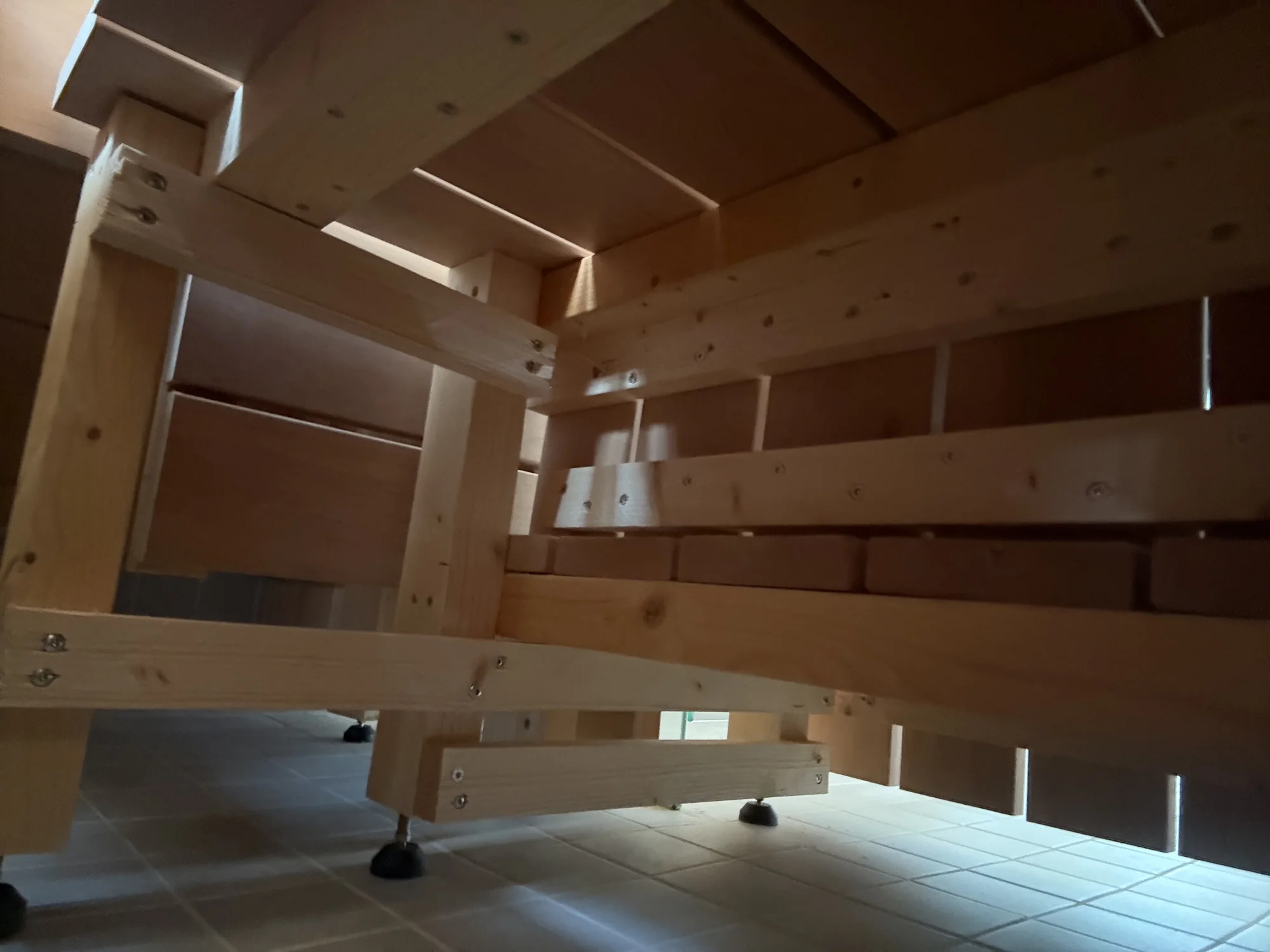 The underside of the sauna benches: softwood posts and rails screwed together, standing on black adjustable feet on a tiled floor