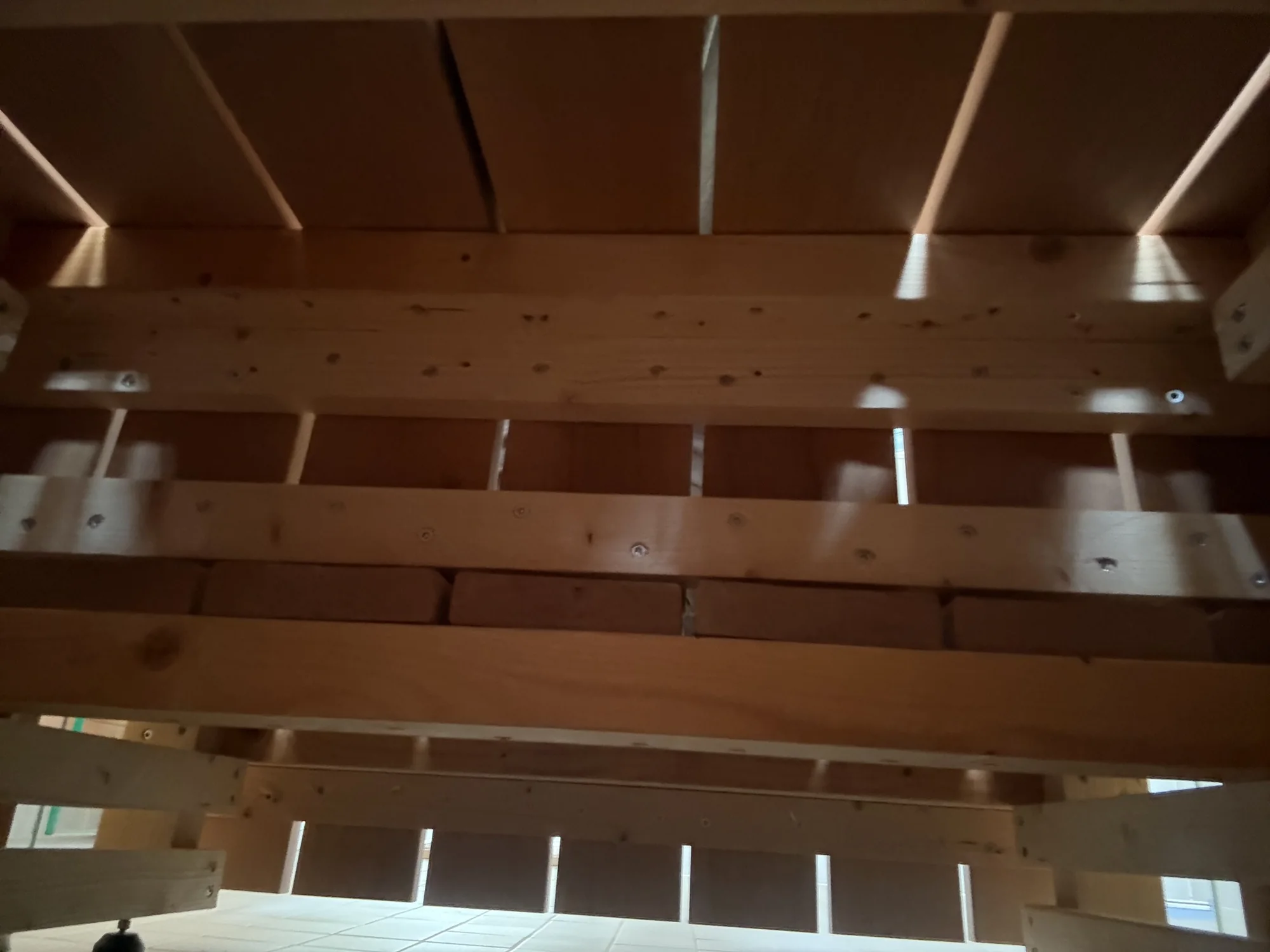 The underside of a sauna bench seen from the tile floor: bench boards with light between them and pale softwood framing members held with screws