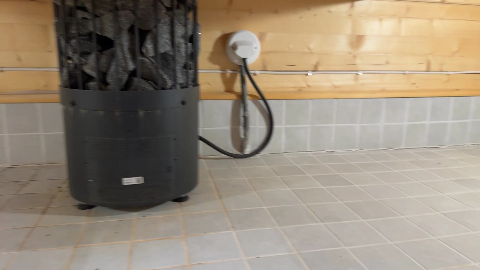The base of the black heater standing on the tiled floor under the platform, with its cable running to a round connection on the wall