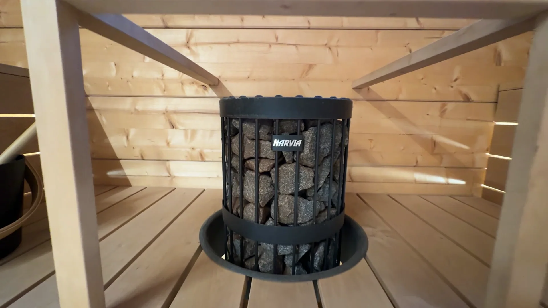 A black cylindrical Harvia heater full of stones rising through a round opening in the sauna platform, with a square wooden guard rail around it