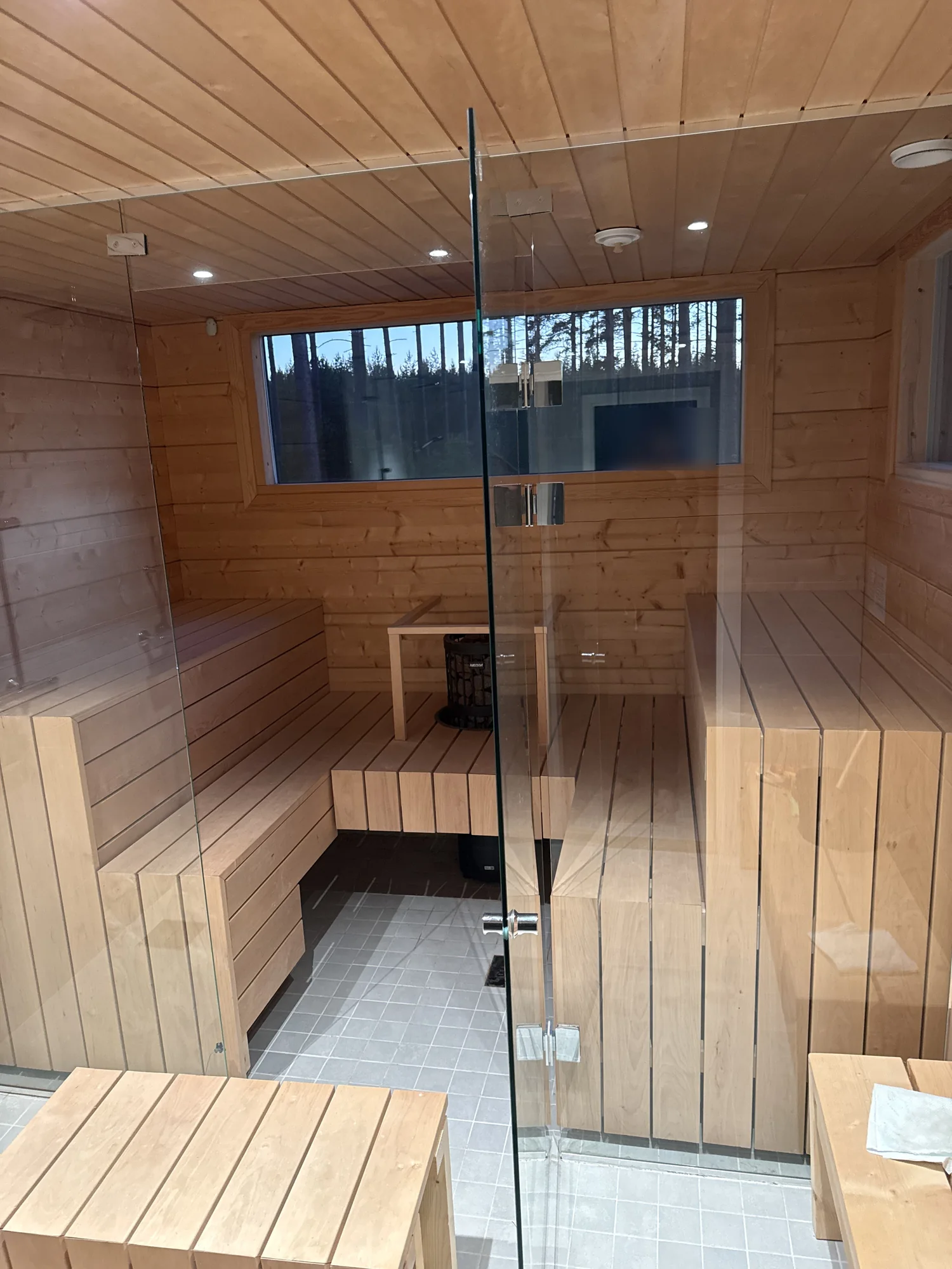 The indoor sauna at Villa Panorama Saimaa seen through its glass front: two benches facing each other, a platform across the back and the heater rising through it behind a wooden guard rail