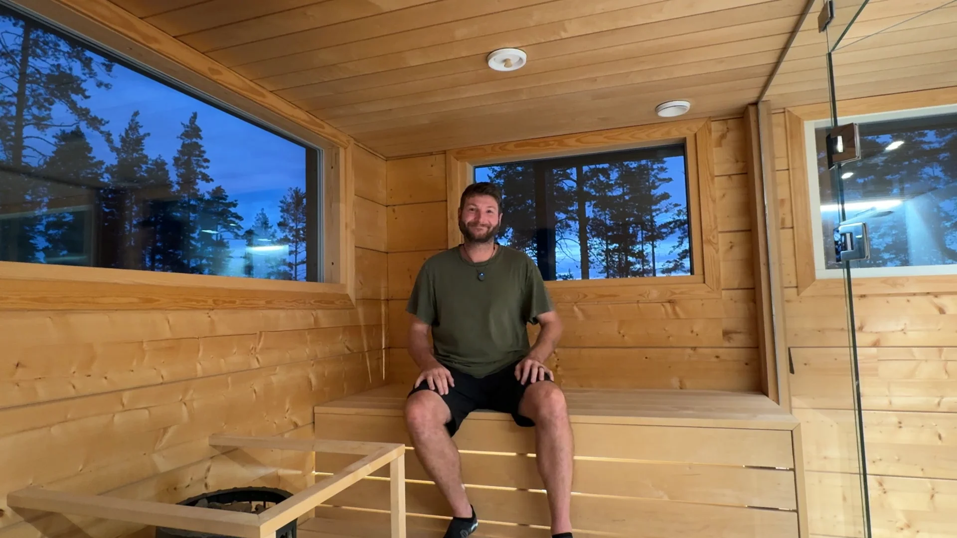 Ben Frizzell smiling on the upper bench of the indoor sauna at Villa Panorama Saimaa at dusk, with pine trees and blue sky in the windows behind him and the heater guard in the corner