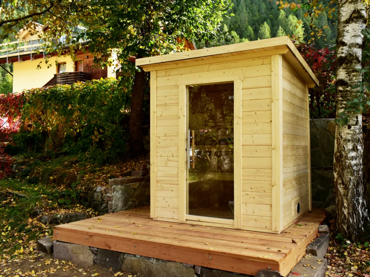 SaunaLife G2 sauna in natural Nordic Spruce with a clear glass door, on a wooden deck in an autumn garden